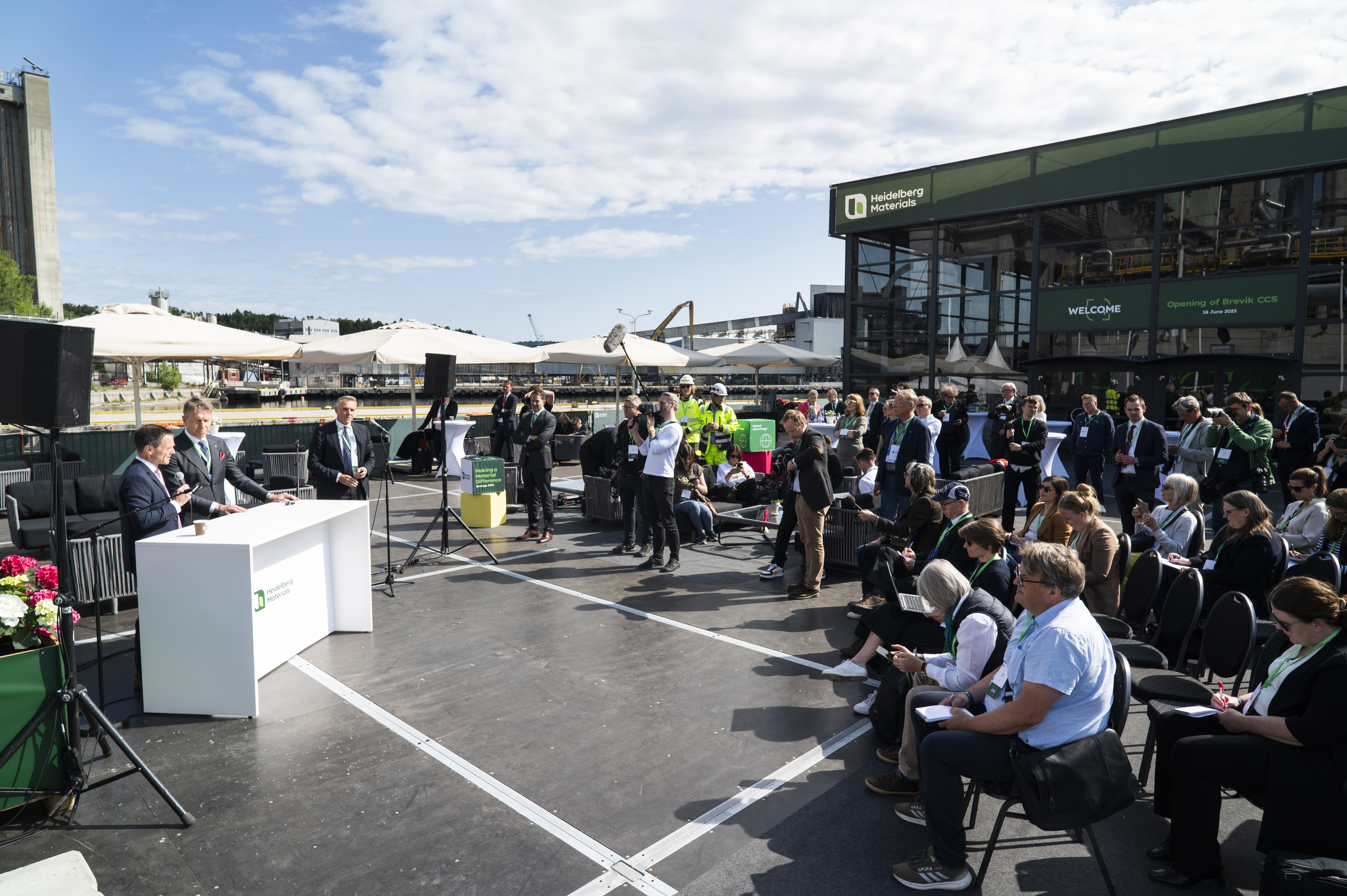 En utomhusscen med en person som håller tal vid ett podium till vänster, omgiven av ett publikhav sittandes till höger. I bakgrunden syns en modern byggnad med gröna dekaler samt parasoller. Himlen är klarblå med vita moln.