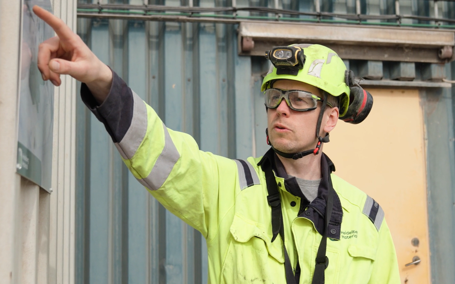 Person i neonfärgad arbetsjacka och hjälm med pannlampa pekar med armen mot något utanför bildens vänstra kant. Bakgrunden visar en industribyggnad med metallväggar och en gul dörr.
