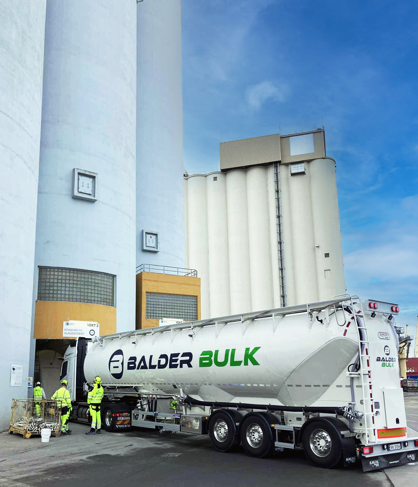 En lastbil från Balder Bulk lossar vid en stor siloanläggning. Fyra personer i gula arbetskläder står intill lastbilen och silobyggnaden. Klarblå himmel i bakgrunden.