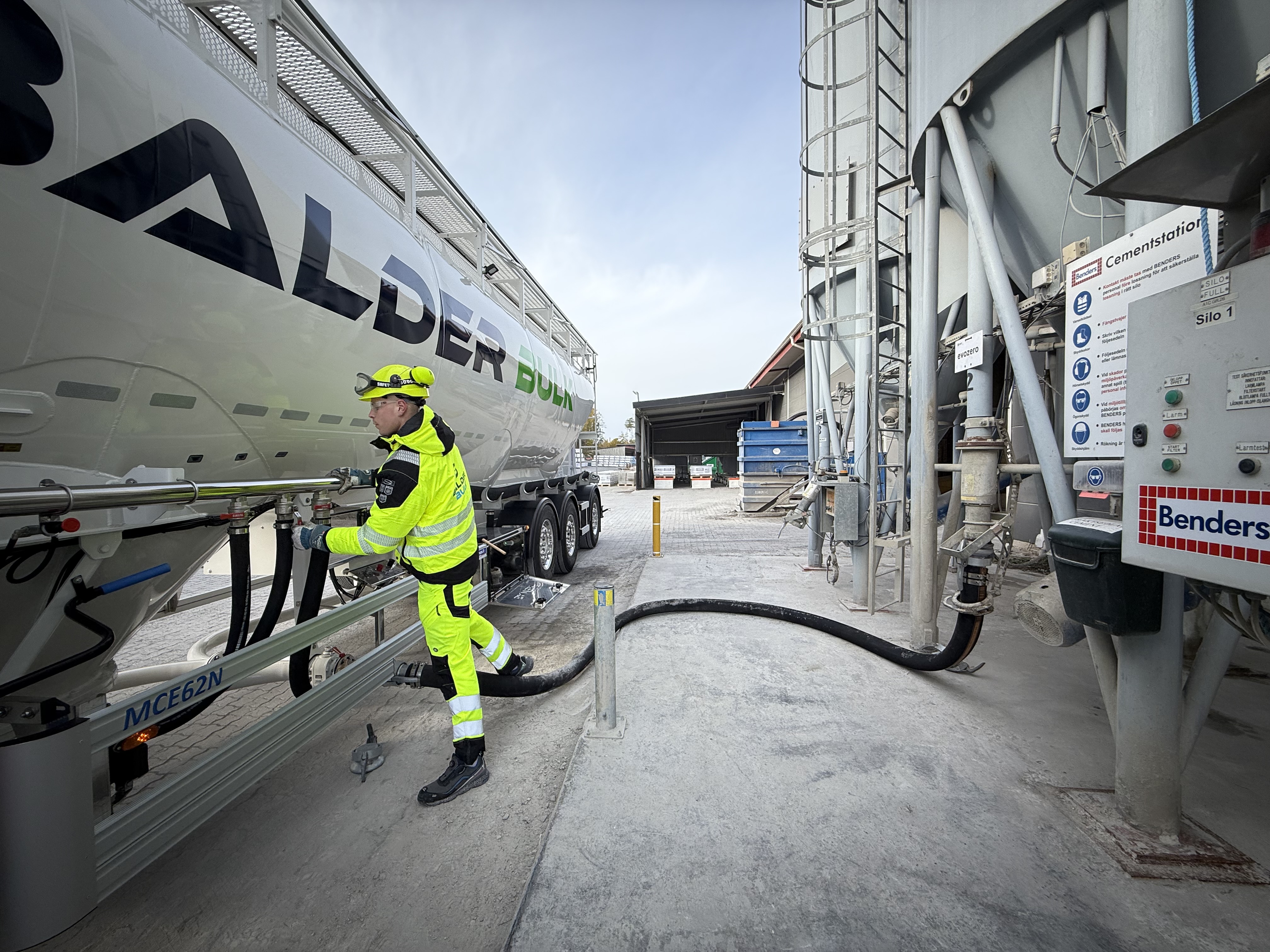 En arbetare i reflexkläder kopplar en slang till en lastbil med företagslogotypen "Balder". Lastbilen står parkerad vid en anläggning med utrustning och en fördelningspanel till höger. Betong- eller materialhanteringsutrustning syns i bakgrunden under en ljus och molnig himmel.