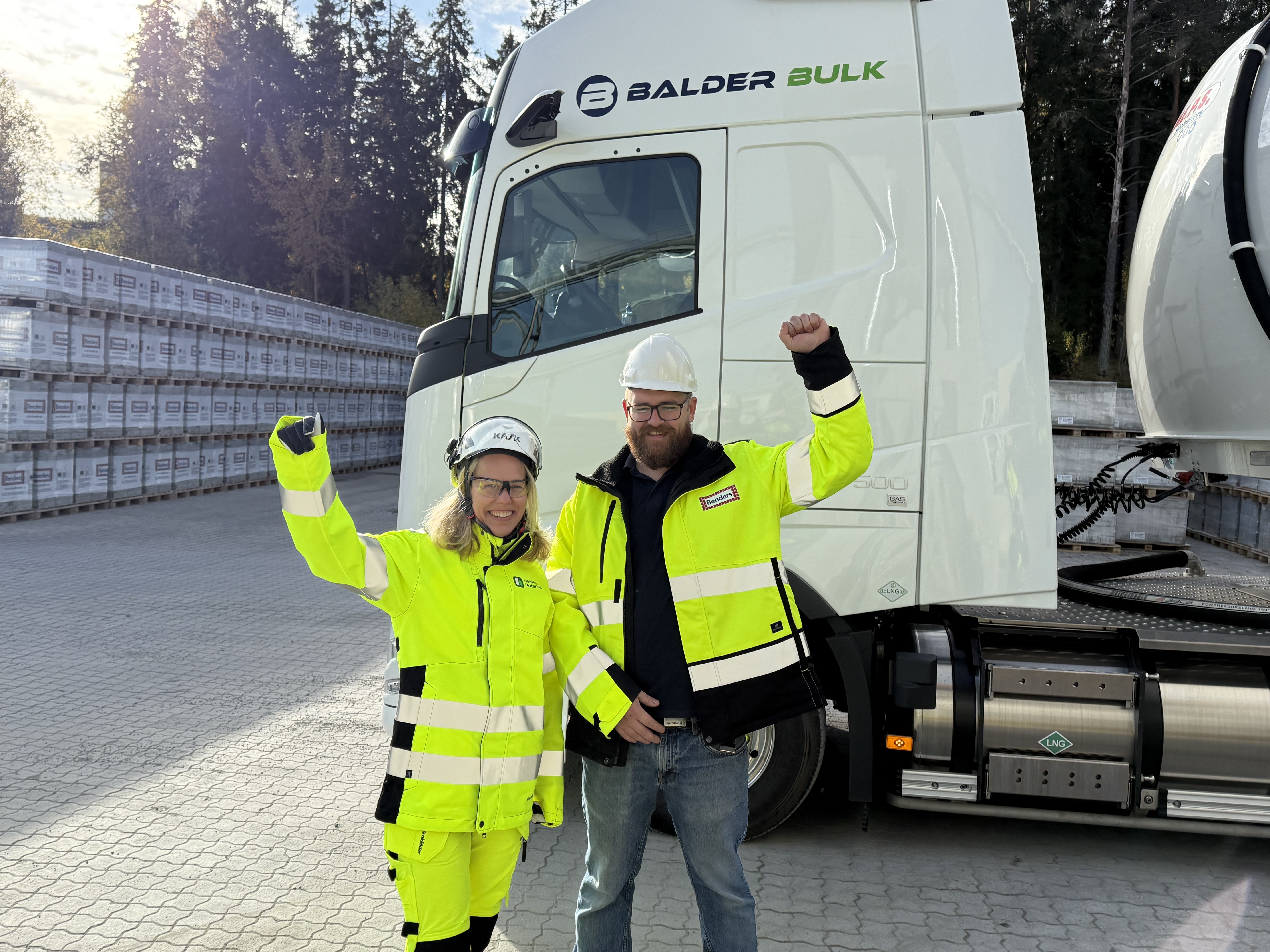 Två personer i reflexkläder och hjälmar står framför en vit lastbil märkt med "Balder Bulk". De håller upp en arm i luften i en segergest. Lastbilen står på en asfalterad plats omgiven av staplade material och träd i bakgrunden.