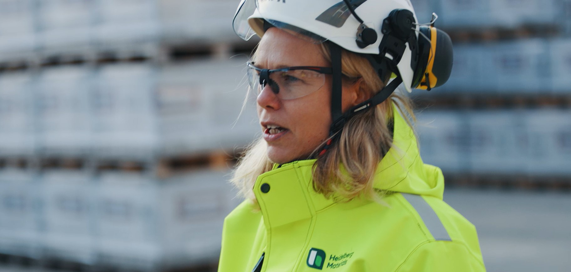 Person i gul skyddsjacka och vit hjälm med hörselskydd, står framför staplade materialpallar utomhus på en arbetsplats.