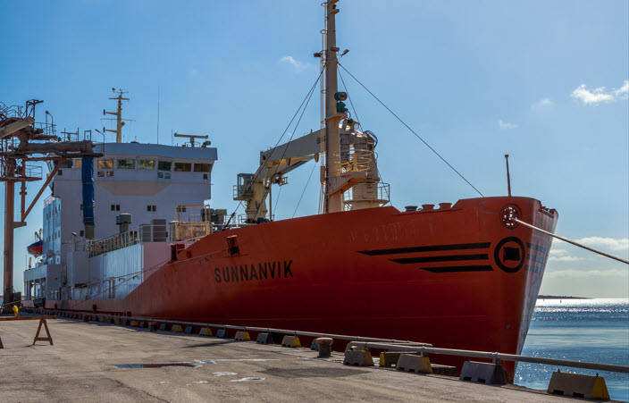 Rött fraktfartyg förtöjt vid en kaj, och solbelyst himmel ovanför.