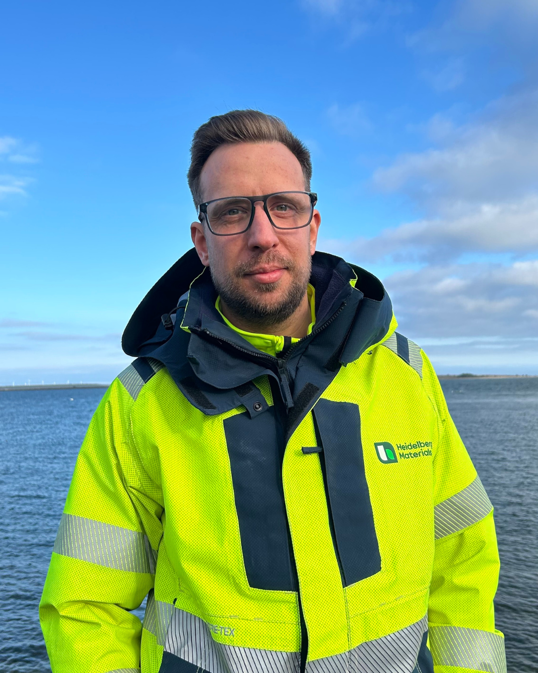 En person klädd i en gul reflexjacka med en mörkblå krage står framför en vattenbakgrund på en solig dag med lätt molnighet.