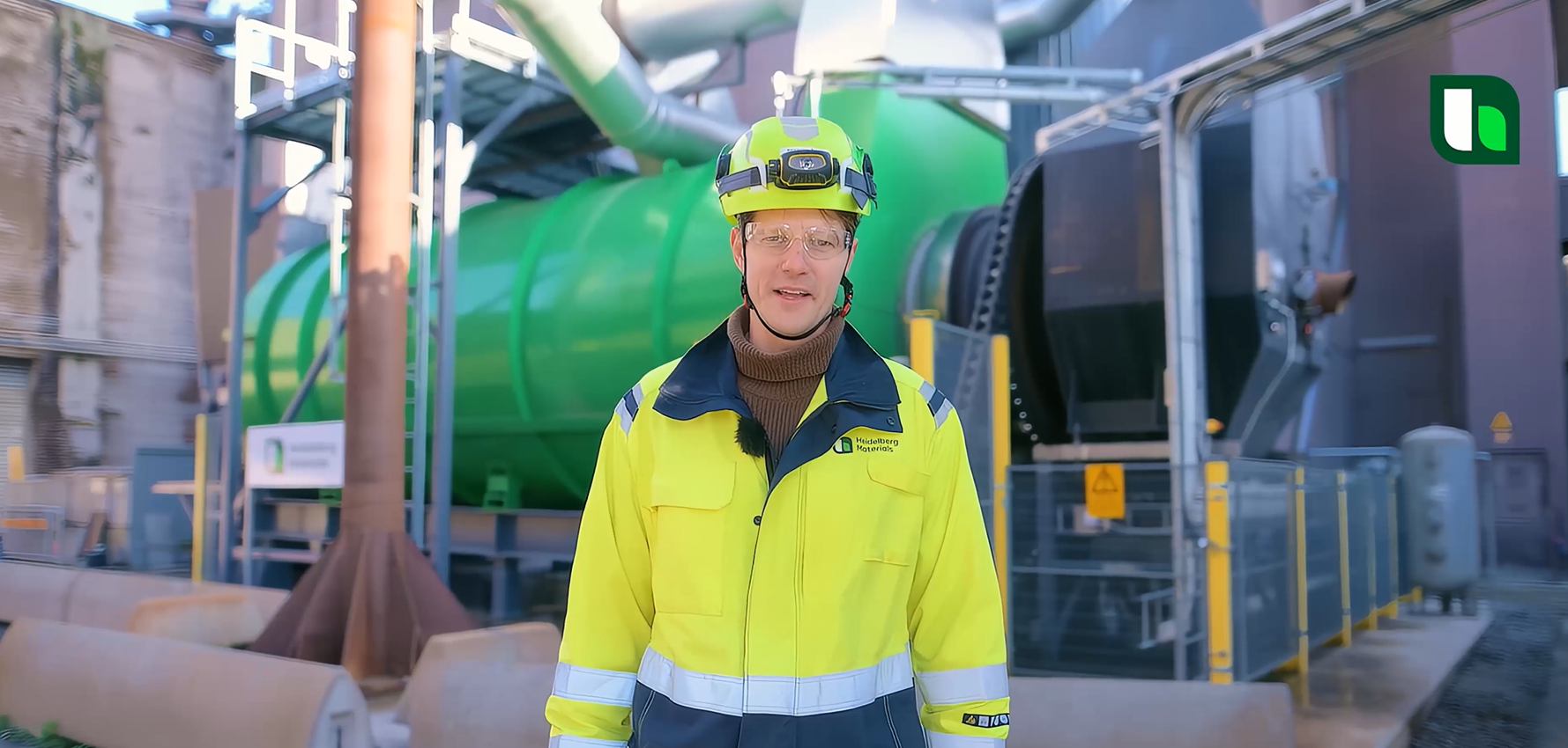 Medarbetare i skyddsutrustning vid en cementanläggning med rör och silo.