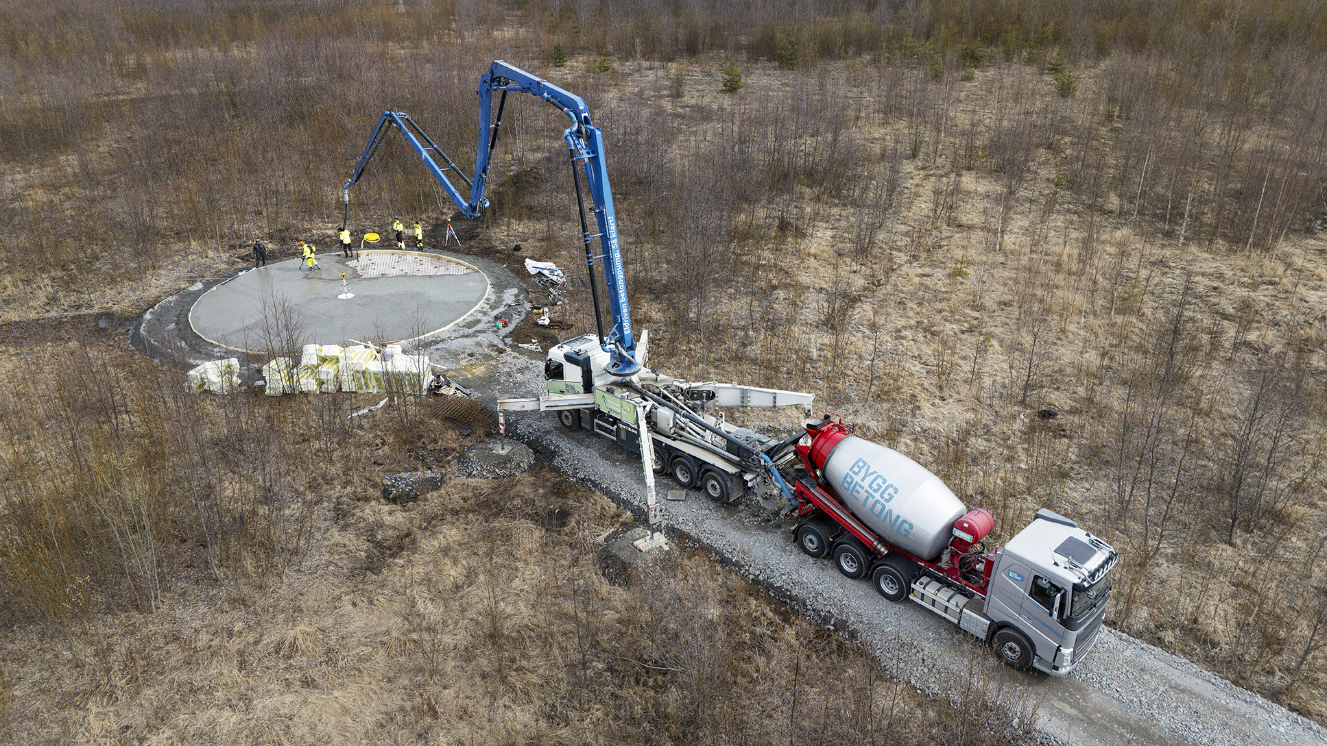 Betongpump och betonglastbil vid byggarbetsplats omgiven av skog.