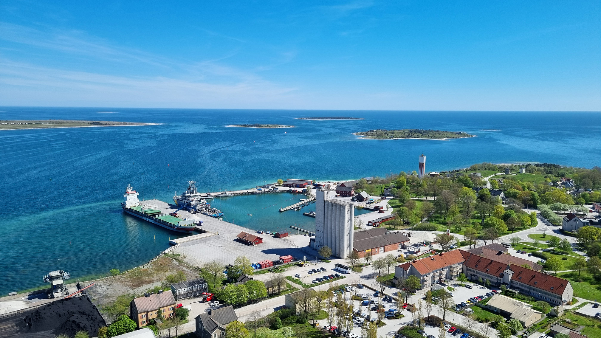 Flygvy över hamnen i Slite med industriell verksamhet, omgiven av vatten och grönska.