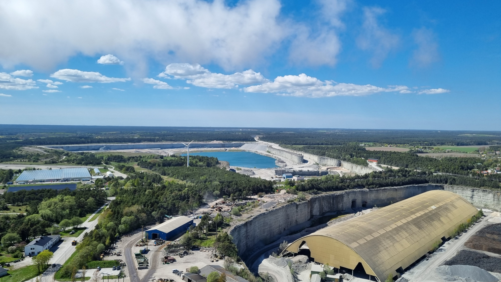 Flygbild av ett industriområde med fabriksbyggnader och en stor stenbrottsanläggning omgiven av skog och vatten.