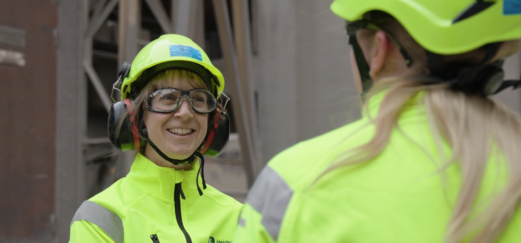 Två personer i varselkläder och hjälmar samtalar på en industriell arbetsplats.