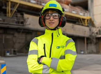 En person i skyddskläder och hjälm står framför en industribyggnad.