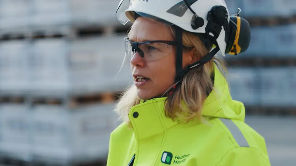 Person i gul skyddsjacka och vit hjälm med hörselskydd, står framför staplade materialpallar utomhus på en arbetsplats.