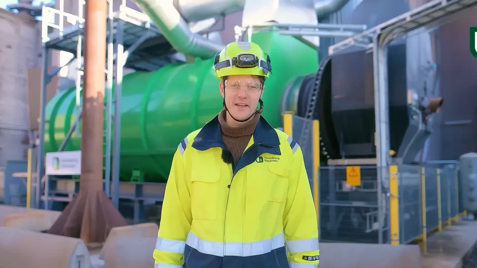 Medarbetare i skyddsutrustning vid en cementanläggning med rör och silo.