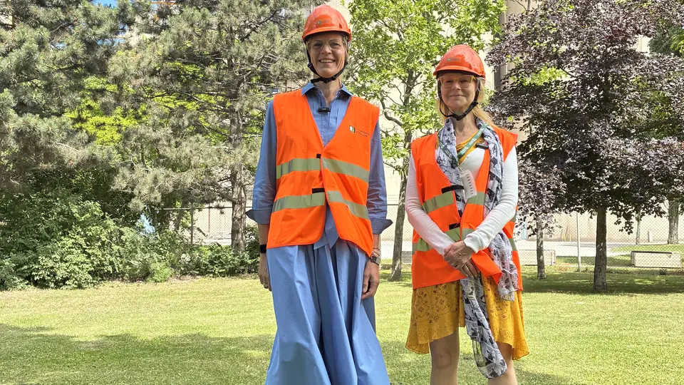 Två personer med orangea säkerhetsvästar och skyddshjälmar står utomhus på en gräsmatta.