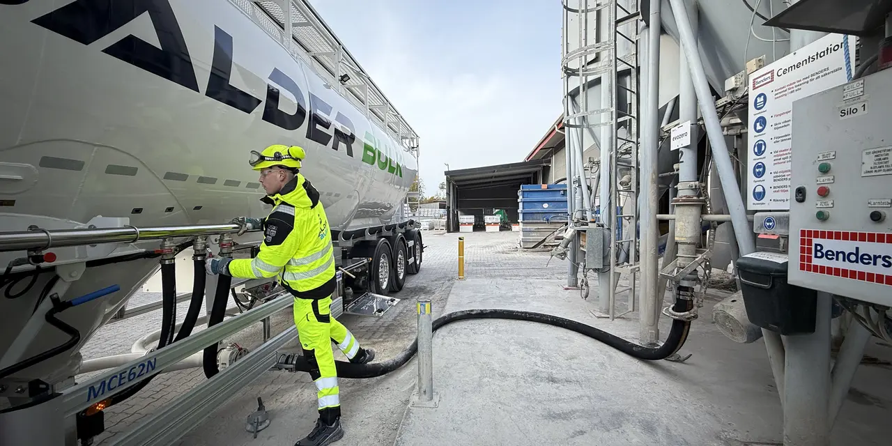 En arbetare i reflexkläder kopplar en slang till en lastbil med företagslogotypen "Balder". Lastbilen står parkerad vid en anläggning med utrustning och en fördelningspanel till höger. Betong- eller materialhanteringsutrustning syns i bakgrunden under en ljus och molnig himmel.