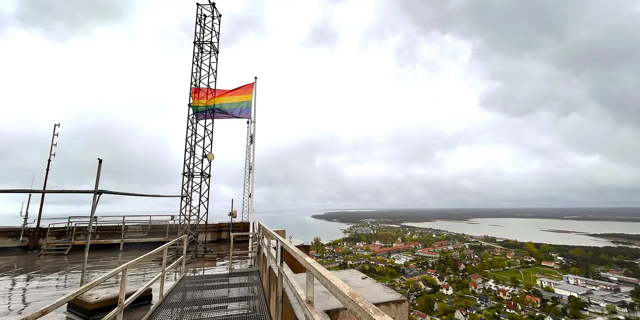 En regnbågsflagga vajar på Slite cementfabrik med utsikt över ett landskap och vatten i bakgrunden.