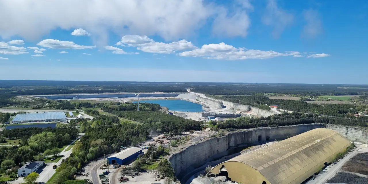 Flygbild av ett industriområde med fabriksbyggnader och en stor stenbrottsanläggning omgiven av skog och vatten.