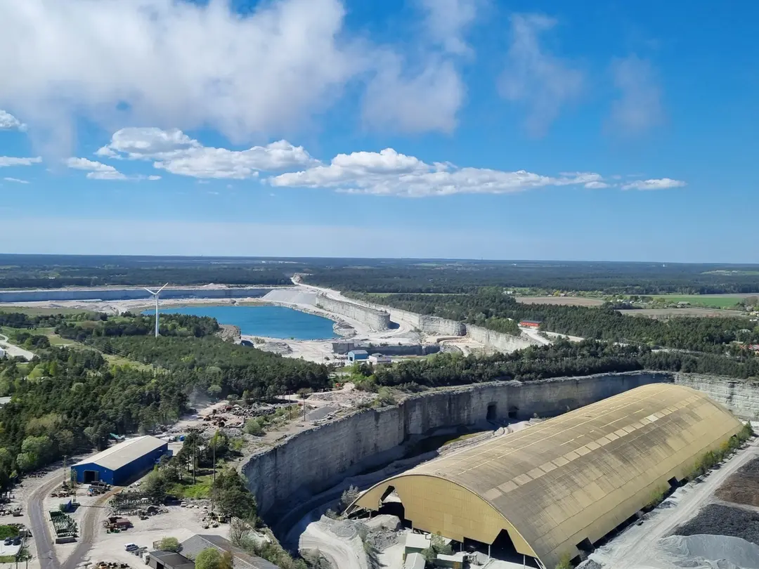 Flygbild av ett industriområde med fabriksbyggnader och en stor stenbrottsanläggning omgiven av skog och vatten.