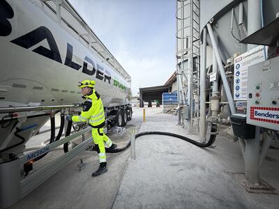 En arbetare i reflexkläder kopplar en slang till en lastbil med företagslogotypen "Balder". Lastbilen står parkerad vid en anläggning med utrustning och en fördelningspanel till höger. Betong- eller materialhanteringsutrustning syns i bakgrunden under en ljus och molnig himmel.